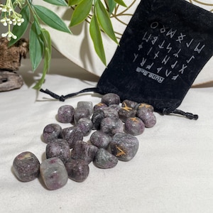 May include: A collection of tumbled stones in shades of purple and gray, scattered on a white surface. A black velvet bag with white runes and the text "Rune Sets" is partially visible. Green leaves and white flowers are in the background.