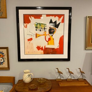 May include: Framed artwork depicting an abstract figure playing a trumpet, with a black crown and a red and white backdrop. The artwork hangs on a light grey wall, above a wooden cabinet with decorative items.
