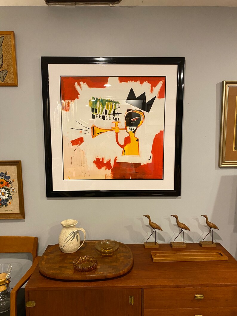 May include: Framed artwork depicting an abstract figure playing a trumpet, with a black crown and a red and white backdrop. The artwork hangs on a light grey wall, above a wooden cabinet with decorative items.