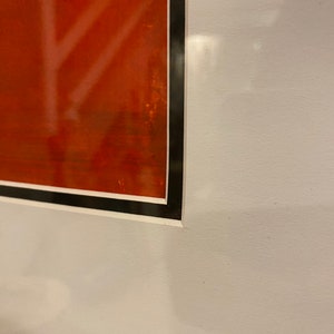 May include: Framed artwork with a vivid red background and white, abstract lines. The artwork is enclosed in a black frame with a white border, creating a striking contrast. The piece is mounted on a white wall.