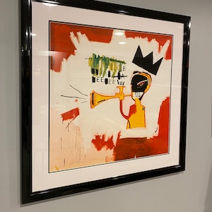 May include: Framed art print depicting an abstract figure playing a trumpet, wearing a black crown. The artwork has a white background with red and green brushstrokes. The print is encased in a black frame.