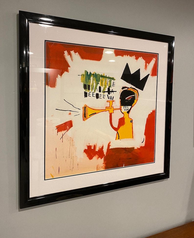 May include: Framed art print depicting an abstract figure playing a trumpet, wearing a black crown. The artwork has a white background with red and green brushstrokes. The print is encased in a black frame.