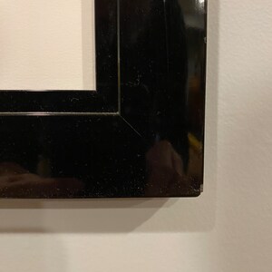 May include: Close-up of a black, glossy picture frame. The frame has a rectangular shape with a beveled edge and a shiny finish. The frame is set against a white wall.