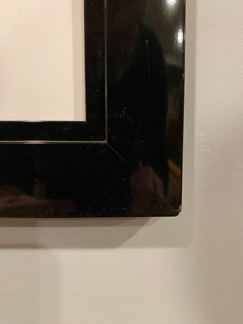 May include: Close-up of a black, glossy picture frame. The frame has a rectangular shape with a beveled edge and a shiny finish. The frame is set against a white wall.
