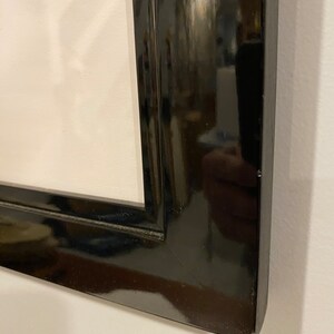 May include: Close-up of a glossy black picture frame. The frame has a rectangular shape with a beveled edge. The frame is positioned against a white wall, reflecting the surrounding environment.