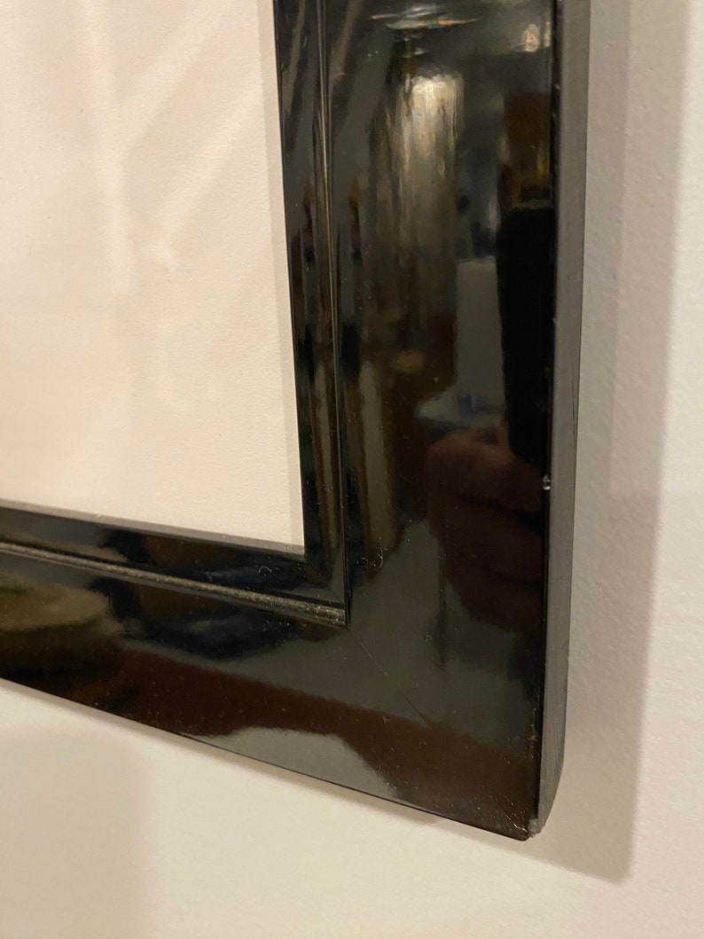 May include: Close-up of a glossy black picture frame. The frame has a rectangular shape with a beveled edge. The frame is positioned against a white wall, reflecting the surrounding environment.
