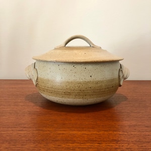 May include: A speckled beige ceramic lidded casserole dish with a curved handle on the lid. The dish has a band of brown around the middle and two decorative handles on the sides. The dish is sitting on a wooden surface.