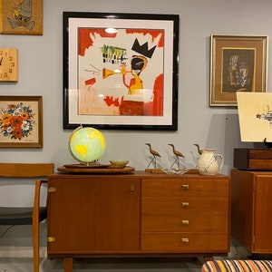 May include: A teak mid-century cabinet with four drawers and a door, featuring brass handles. A globe, decorative birds, and framed art, including a piece with a crown, sit on top. Other framed artworks are displayed on the grey wall.