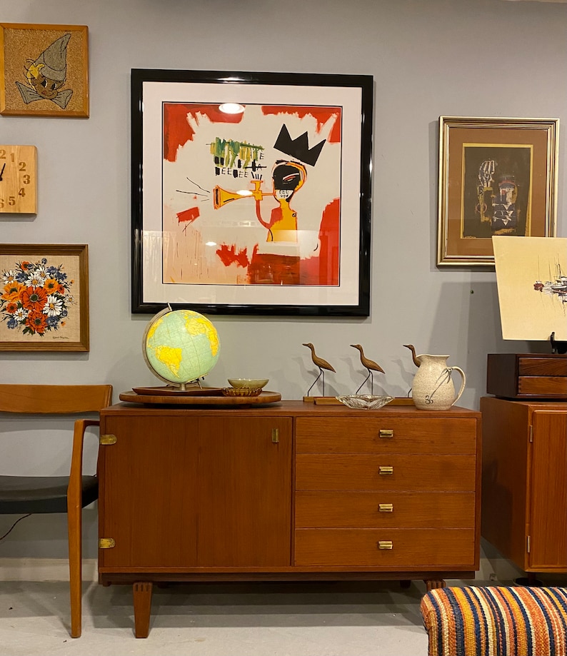 May include: A teak mid-century cabinet with four drawers and a door, featuring brass handles. A globe, decorative birds, and framed art, including a piece with a crown, sit on top. Other framed artworks are displayed on the grey wall.