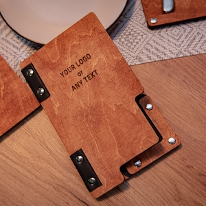 May include: A wooden menu cover with black leather accents and silver rivets. The cover is engraved with the text "YOUR LOGO or ANY TEXT".