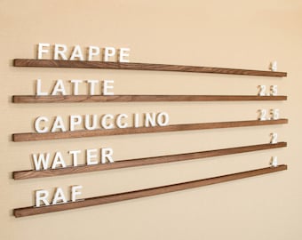 Könnte beinhalten: Eine Holztafel mit weißen Buchstaben, die die Getränkeoptionen angeben: Frappe, Latte, Cappuccino, Wasser und Raf. Die Preise sind neben jedem Getränk aufgeführt.
