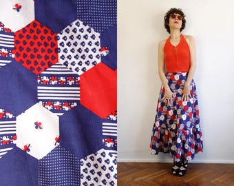 May include: A long skirt featuring a patchwork-style hexagonal pattern in red, white, and navy blue. The fabric displays various geometric motifs including stripes, dots, and small floral shapes within the hexagons. The skirt has a ruffled hemline.