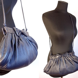 May include: A navy blue pleated crossbody bag with a bow detail. The bag has a long, thin strap and is displayed on a black mannequin. The bag's design features vertical pleats and a decorative bow on the front.
