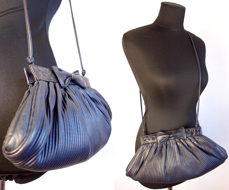 May include: A navy blue pleated crossbody bag with a bow detail. The bag has a long, thin strap and is displayed on a black mannequin. The bag's design features vertical pleats and a decorative bow on the front.