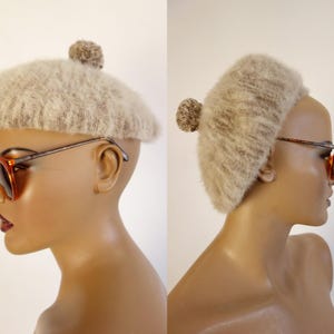 May include: A beige, fuzzy beret with a small pom-pom on top. The hat is displayed on a mannequin head, paired with orange-tinted sunglasses with a brown frame. The beret has a soft, textured appearance.