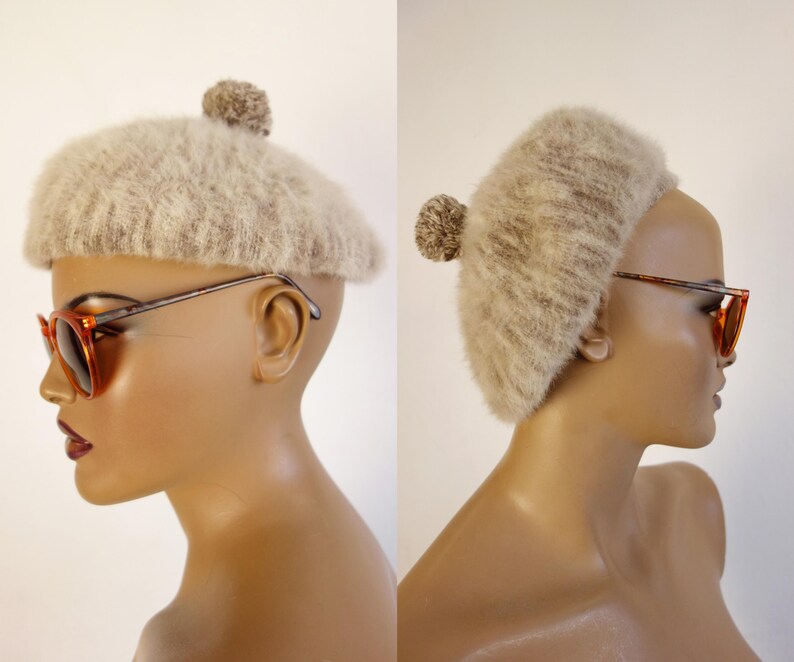 May include: A beige, fuzzy beret with a small pom-pom on top. The hat is displayed on a mannequin head, paired with orange-tinted sunglasses with a brown frame. The beret has a soft, textured appearance.