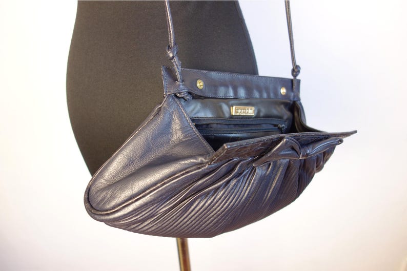 May include: A dark navy blue leather crossbody bag with a pleated design. The bag has a flap closure and a long strap. The interior is visible, revealing a zippered pocket and a label that reads "SPORTE".