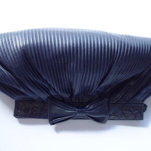 May include: Navy blue clutch with a pleated design and a bow detail. The clutch has a rectangular shape with a textured, embossed band at the base. The bow is centered on the front of the clutch.
