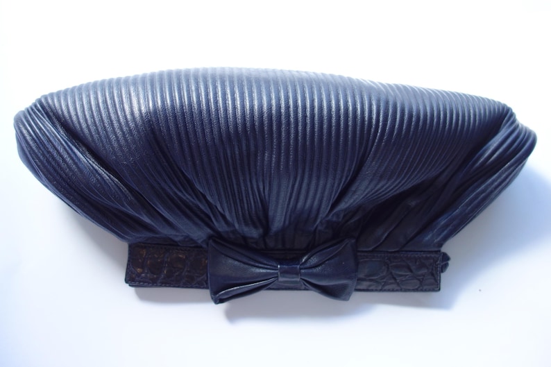 May include: Navy blue clutch with a pleated design and a bow detail. The clutch has a rectangular shape with a textured, embossed band at the base. The bow is centered on the front of the clutch.