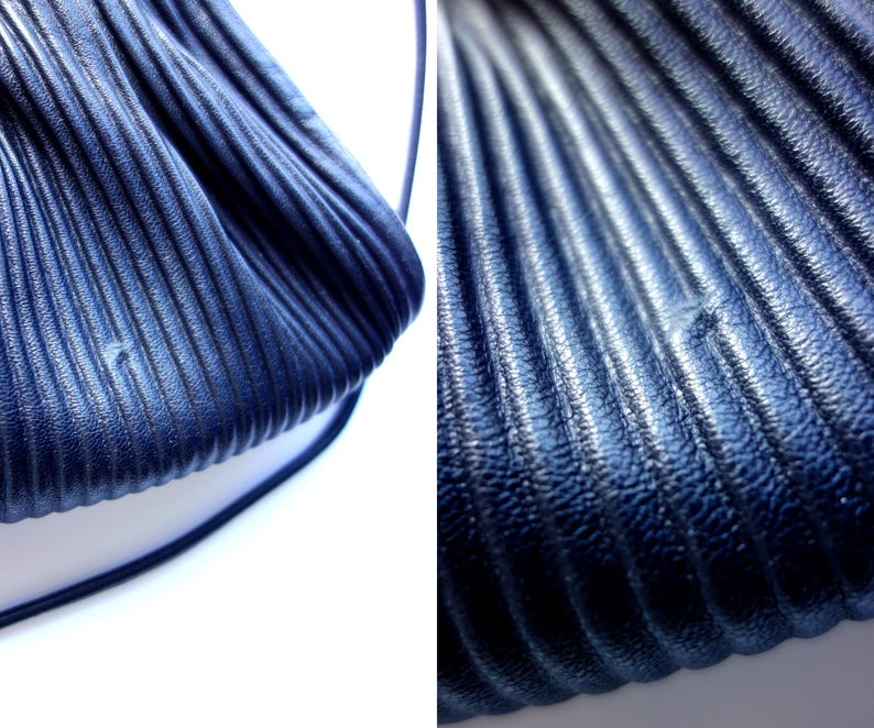 May include: Close-up of a navy blue, pleated handbag. The bag has a textured, ridged surface and a thin, matching strap. The image highlights the bag's design and material, showcasing its unique texture and color.