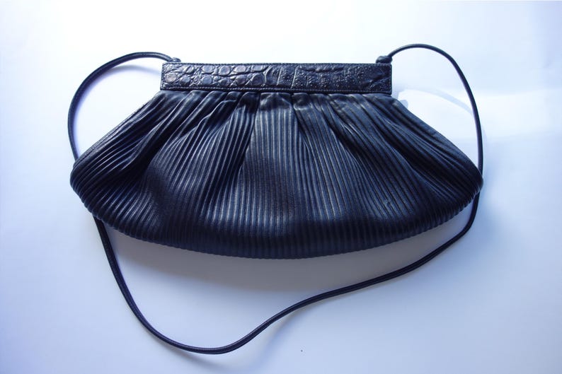 May include: Black pleated handbag with a long strap. The top of the bag has a textured, crocodile-like design. The bag's body is made of a pleated material, creating a textured effect. The strap is thin and appears to be made of the same material as the bag.