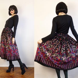 May include: A long, pleated skirt featuring a black base with a vibrant, multi-coloured floral and paisley pattern. The hem is accented with a vertical striped border in shades of red, blue, and purple. The skirt is paired with a black long-sleeved top.