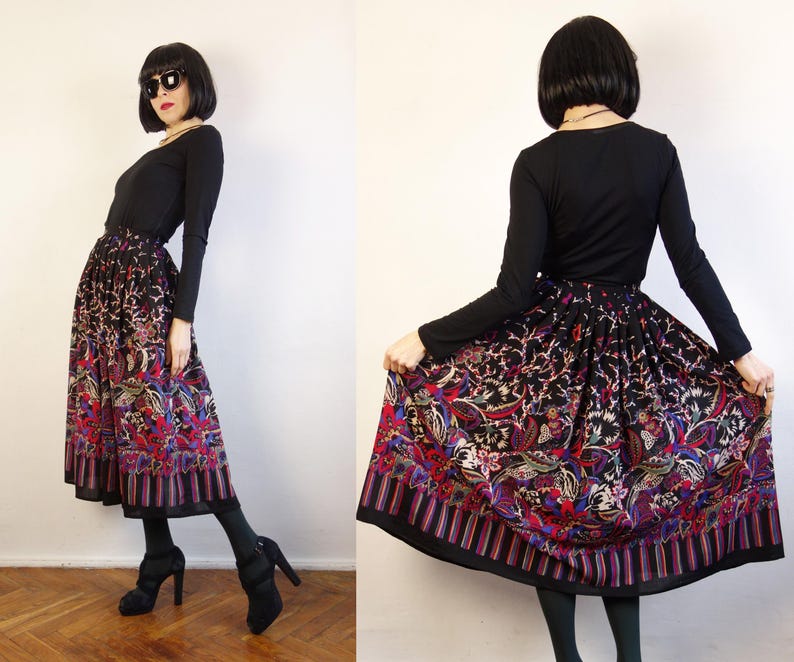 May include: A long, pleated skirt featuring a black base with a vibrant, multi-coloured floral and paisley pattern. The hem is accented with a vertical striped border in shades of red, blue, and purple. The skirt is paired with a black long-sleeved top.
