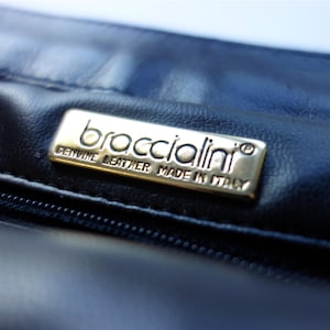 May include: Close-up of a dark blue leather handbag. A rectangular, gold-colored metal tag is affixed to the leather, with the brand name "broccialini" and the text "GENUINE LEATHER MADE IN ITALY" engraved on it.
