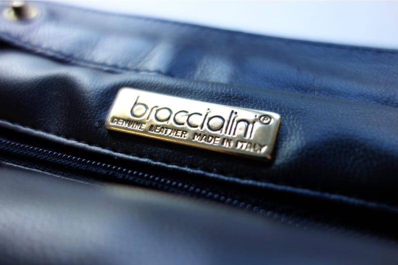 May include: Close-up of a dark blue leather handbag. A rectangular, gold-colored metal tag is affixed to the leather, with the brand name "broccialini" and the text "GENUINE LEATHER MADE IN ITALY" engraved on it.