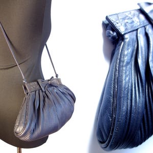 May include: A navy blue, pleated leather crossbody bag with a bow detail on the flap. The bag has a long, thin strap and a rounded shape. The bag is displayed on a black mannequin.