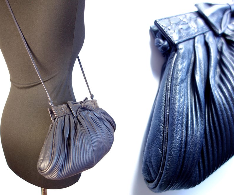 May include: A navy blue, pleated leather crossbody bag with a bow detail on the flap. The bag has a long, thin strap and a rounded shape. The bag is displayed on a black mannequin.