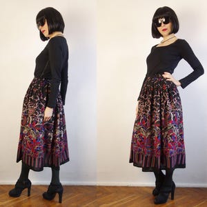 May include: A long-sleeved black top paired with a mid-calf length skirt featuring a vibrant, multi-coloured floral and abstract pattern on a black background, finished with a vertical striped border at the hemline and a gathered, full silhouette.