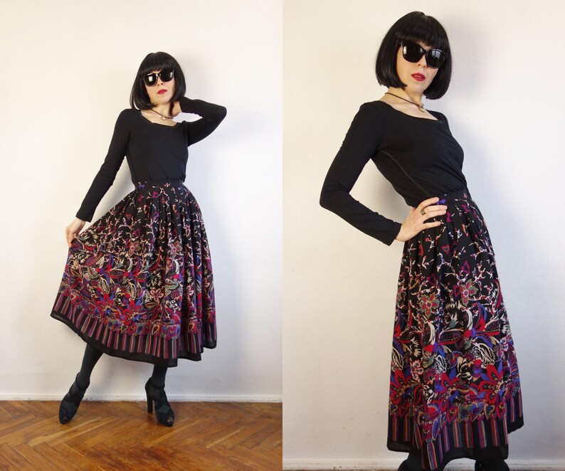 May include: A long-sleeved black top paired with a high-waisted, pleated midi skirt featuring a vibrant, multi-coloured floral and paisley pattern on a black background, finished with a striped border at the hemline.