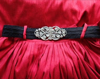May include: A bright red pleated skirt featuring a black textured belt with a silver-toned metal buckle. The buckle displays a symmetrical floral design with intricate leaf and petal details, secured by two red belt loops on the waistband.