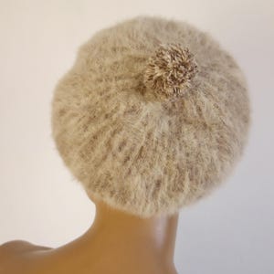 May include: A beige beret with a fluffy texture and a small pom-pom on top. The hat is made of a soft, fuzzy material and is designed to be worn on the head. The beret is a neutral color, making it versatile for various outfits.