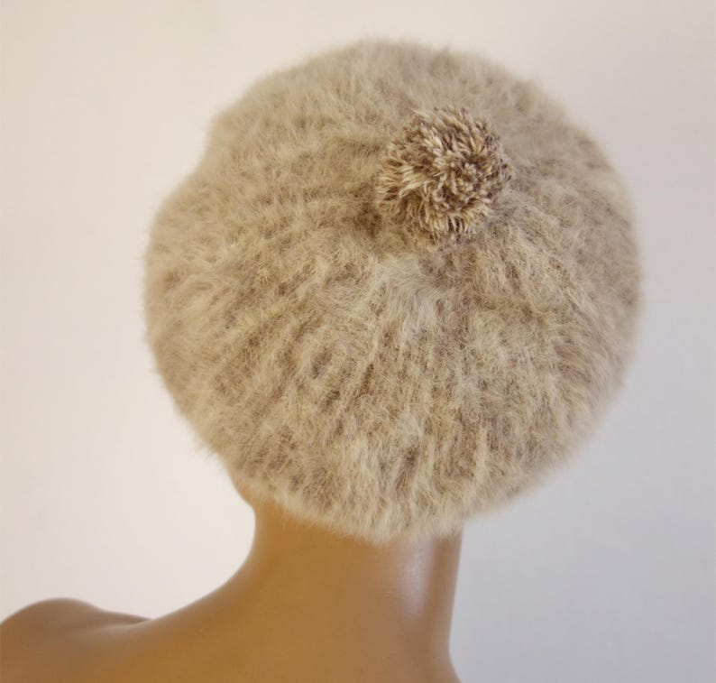 May include: A beige beret with a fluffy texture and a small pom-pom on top. The hat is made of a soft, fuzzy material and is designed to be worn on the head. The beret is a neutral color, making it versatile for various outfits.