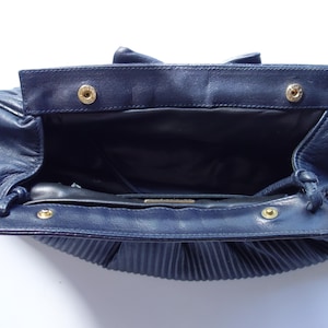 May include: Open navy blue leather handbag with a pleated bottom. The bag has a rectangular flap with two gold-colored snap closures. The interior is black, and the bag is open, revealing the inside.