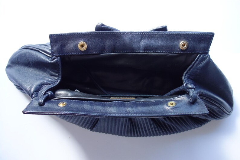 May include: Open navy blue leather handbag with a pleated bottom. The bag has a rectangular flap with two gold-colored snap closures. The interior is black, and the bag is open, revealing the inside.