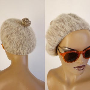May include: A light brown fuzzy beret with a small pom-pom on top. The beret is displayed on a mannequin head, paired with orange-framed sunglasses. The beret is a stylish accessory.