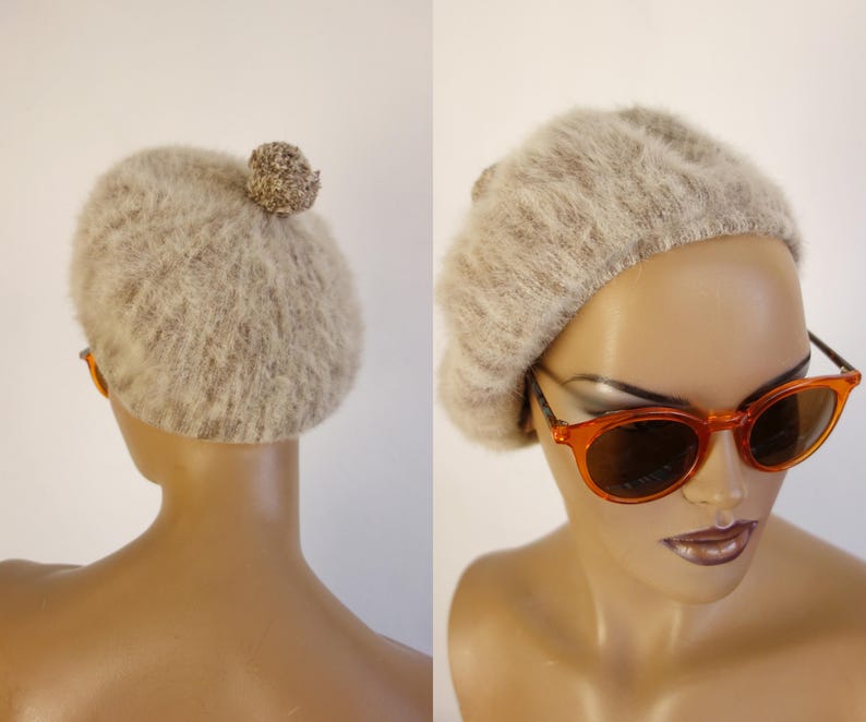 May include: A light brown fuzzy beret with a small pom-pom on top. The beret is displayed on a mannequin head, paired with orange-framed sunglasses. The beret is a stylish accessory.