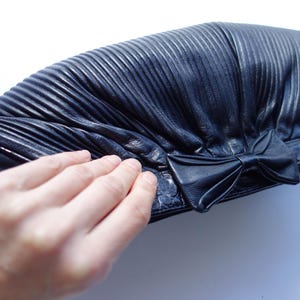 May include: A black leather clutch with a pleated design and a bow detail. The clutch has a textured surface and a rectangular shape. The image shows the clutch on a white background.