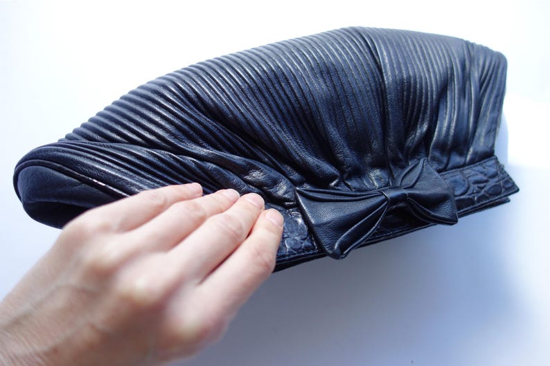 May include: A black leather clutch with a pleated design and a bow detail. The clutch has a textured surface and a rectangular shape. The image shows the clutch on a white background.