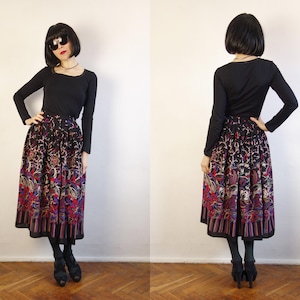 May include: A long, pleated skirt featuring a dark base with a vibrant, multi-coloured floral and paisley pattern, paired with a solid black long-sleeved top. The skirt has a wide, striped border at the hem and falls below the knee.