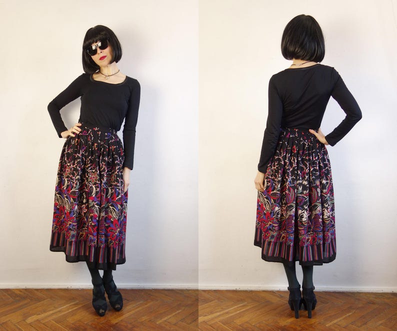 May include: A long, pleated skirt featuring a dark base with a vibrant, multi-coloured floral and paisley pattern, paired with a solid black long-sleeved top. The skirt has a wide, striped border at the hem and falls below the knee.