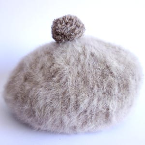 May include: A light brown, fluffy beret with a small, textured pom-pom on top. The beret is made of a soft, fuzzy material and is set against a white background. This hat is a stylish accessory for cold weather.