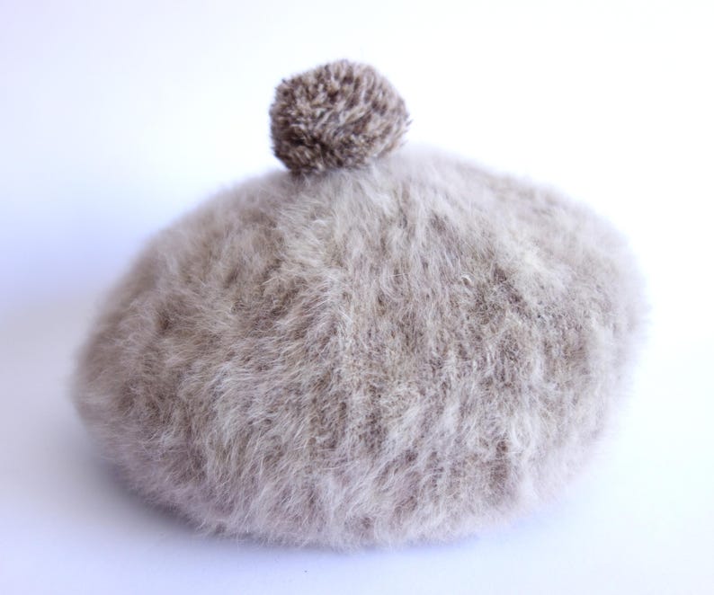 May include: A light brown, fluffy beret with a small, textured pom-pom on top. The beret is made of a soft, fuzzy material and is set against a white background. This hat is a stylish accessory for cold weather.
