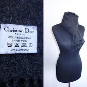May include: A dark gray lambswool scarf with fringe, draped on a black mannequin. A white tag with the text "Christian Dior PARIS" and care instructions is visible. The scarf is made of 100% laine d'agneau.