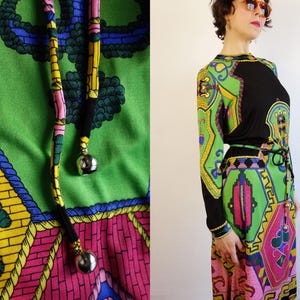 Könnte beinhalten: Ein Vintage-Maxikleid mit einem lebendigen, geometrischen Muster in Grün, Schwarz, Rosa, Gelb und Blau. Das Kleid hat lange Ärmel, ein schwarzes Oberteil und einen passenden Gürtel. Der Kordelzug hat silberne Metallkugelenden.