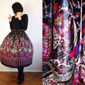 May include: A full-length, high-waisted skirt featuring a vibrant, multi-coloured floral and paisley pattern on a black background. The skirt has a voluminous, pleated silhouette that extends to the mid-calf, paired with a solid black long-sleeved top.