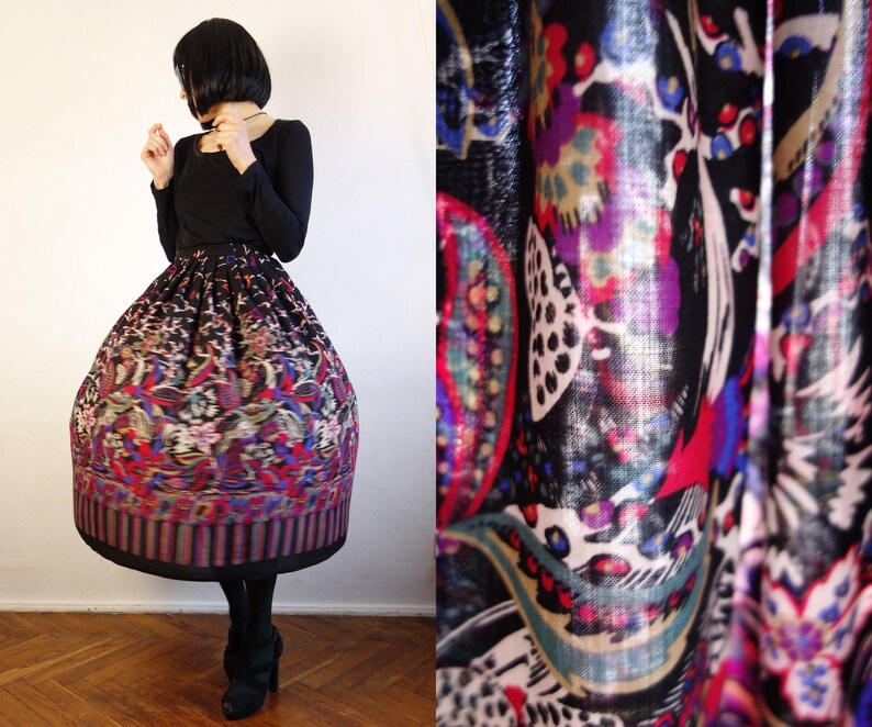 May include: A full-length, high-waisted skirt featuring a vibrant, multi-coloured floral and paisley pattern on a black background. The skirt has a voluminous, pleated silhouette that extends to the mid-calf, paired with a solid black long-sleeved top.